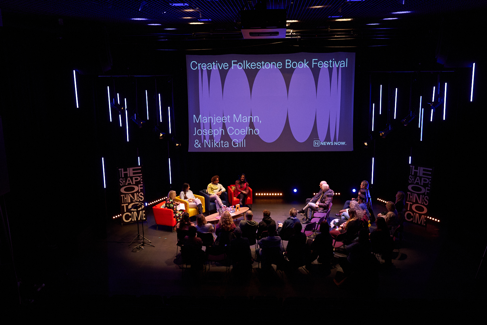 Overhead view of the Book Festival stage: a panel on coloured armchairs below a large screen showing the festival's stretched oval identity, with 'The Shape of Things to Come' banners either side